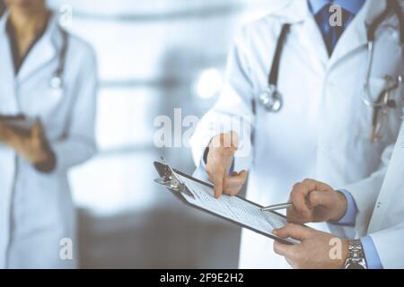 Unbekannte Ärzte, zwei Männer und eine Frau, diskutieren medizinische Untersuchungsresoults, während sie im Krankenhaus-Büro stehen. Ärzte, die Klemmbretter zum Auffüllen verwenden Stockfoto