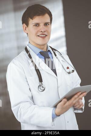Ein junger, freundlicher Arzt überprüft einige Informationen auf seinem Computer-Tablet. Porträt eines Facharztes bei der Arbeit in einer Klinik. Medizinkonzept Stockfoto
