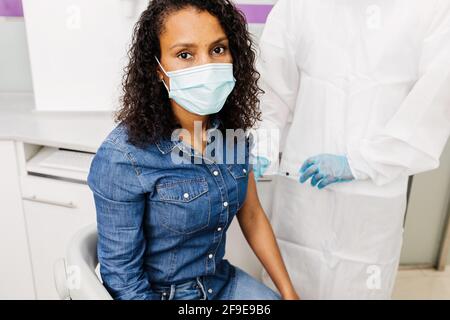 Kurzgeschnittenes, nicht erkennbares medizinisches Fachpersonal mit Schutzkleidung, Latexhandschuhen und Gesichtsschutz, das afroamerikanische weibliche Patienten impft Stockfoto