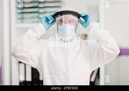 Positiver männlicher Arzt mit Latexhandschuhen und weißer Uniform-Putting Auf Gesichtsschild Schutzmaske stehen in modernen Arztpraxis Und den Kamerun betrachten Stockfoto