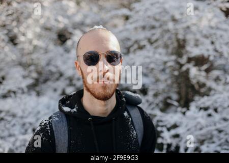 Bärtiger männlicher Rucksacktourist, der während der Kamera gegen verschneite Bäume schaut Reise an einem sonnigen Tag Stockfoto