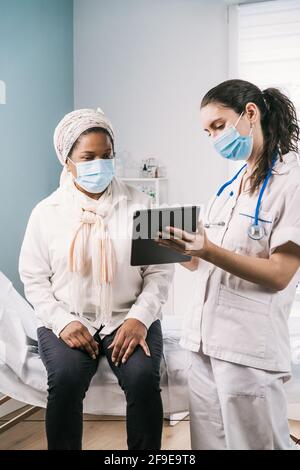 Junge Ärztin in medizinischer Uniform und Stethoskop trägt Gesichtsmaske sprechen und zeigt Ergebnis auf Tablette afroamerikanischen reifen Frau Patien Stockfoto