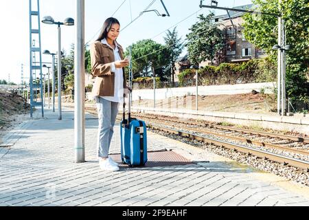 Inhalt Ethnische weibliche Reisende mit Koffer auf Plattform zu stehen Eisenbahnsuche auf dem Smartphone Stockfoto