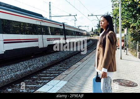 Seitenansicht der asiatischen Reisenden mit Koffer auf dem stand Bahnsteig des Bahnhofs und Blick auf den Zug Stockfoto