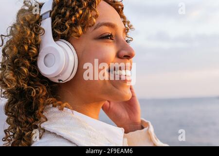 Seitenansicht einer fröhlichen schwarzen Frau in kabellosen Kopfhörern Abends Musik an der Küste Stockfoto