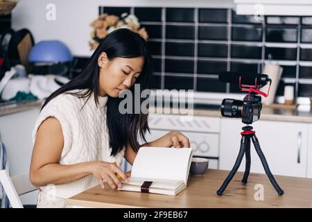 Junge ethnische weibliche Vlogger mit Notebook am Tisch sitzen mit Fotokamera auf Stativ in der Küche Stockfoto