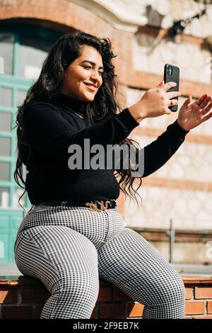 Fröhliche plus size weiblich in trendigen Kleidern sitzen auf Zaun Und Selbstaufnahmen mit dem Smartphone am sonnigen Tag im Hotel Stadt Stockfoto
