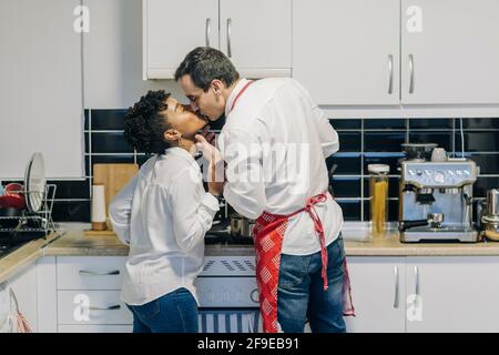 Seitenansicht eines verliebten multiethnischen Paares, das im Stehen küsst In Küche und Kochen Stockfoto