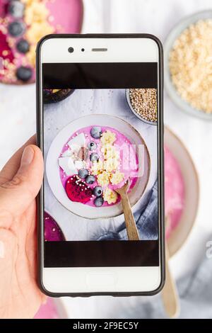 Draufsicht auf Ernte unkenntlich Person demonstriert Fotografie von Delicious Frühstückschüssel mit Smoothie und Obst auf dem Handy-Bildschirm Stockfoto