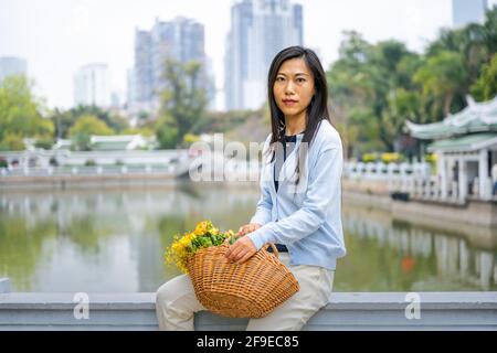 Schöne asiatische Mädchen Porträt in einem Park, während sie neben Weidenkorb mit gelben Blumen sitzt. Stockfoto