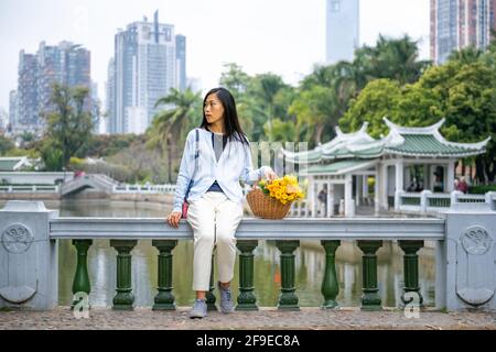 Schöne asiatische Mädchen Porträt in einem Park, während sie neben Weidenkorb mit gelben Blumen sitzt. Stockfoto