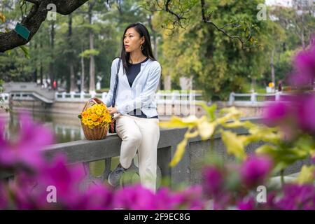 Schöne asiatische Mädchen Porträt in einem Park, während sie neben Weidenkorb mit gelben Blumen sitzt. Stockfoto