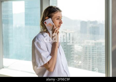 Junge, nachdenkliche Geschäftsfrau, die mit großen Fenstern im Büro steht und einen Anruf auf dem Mobiltelefon führt Stockfoto