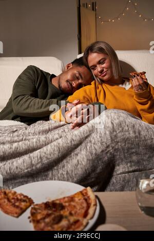 Ein multiethnisches Paar sitzt auf dem Sofa und eine Freundin isst leckere Pizza Während der Freund schläft Stockfoto