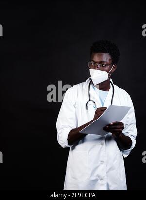 Afroamerikanischer Arzt in Schutzmaske und weißer Uniform Notizen in der Zwischenablage machen, während man auf schwarzen Hintergrund schaut Stockfoto