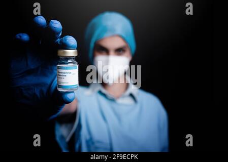 Verschwommener, nicht erkennbarer Sanitäter in Schutzmaske und Latexhandschuhen Zum Kameraglasfläschchen mit Coronavirus-Impfstoff im Stehen Schwarzer Hintergrand Stockfoto