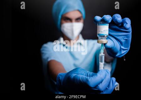 Verschwommener unerkennbarer Sanitäter in Schutzmaske und Latexhandschuhen mit Fläschchen mit Coronavirus-Impfstoff und Spritze auf schwarzem Hintergrund Im Krankenhaus Stockfoto