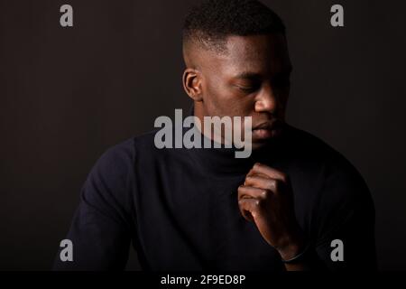 Selbstbewusster junger afroamerikanischer Kerl in schwarzer Kleidung mit der Hand Am Kinn verschlossenen Augen auf schwarzem Hintergrund im Lichtstudio Stockfoto