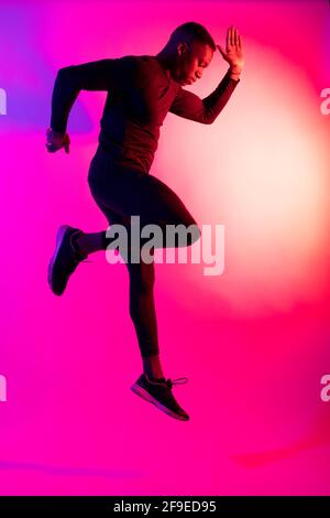 Ganzkörper-Seitenansicht des afroamerikanischen männlichen Athleten in Sportbekleidung springen in lila und rosa Neon Beleuchtung des Studios Stockfoto