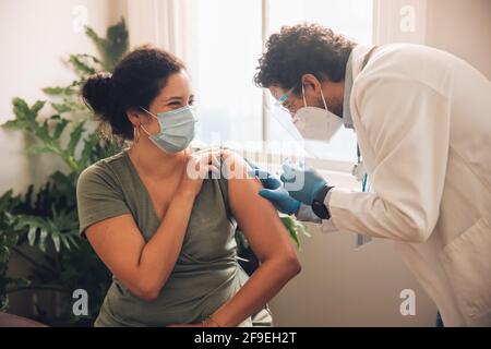 Frau trägt eine schützende Gesichtsmaske und erhält einen Coronavirus-Impfstoff am Arm von einem männlichen Arzt. Frontarbeiterin impft Frau an ihrem Hom Stockfoto