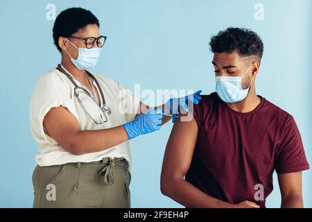 Ärztin mit Gesichtsmaske und Handschuhen, die einem Mann vor blauem Hintergrund eine Impfung verabreichen. Mann wird von einer Ärztin geimpft. Stockfoto