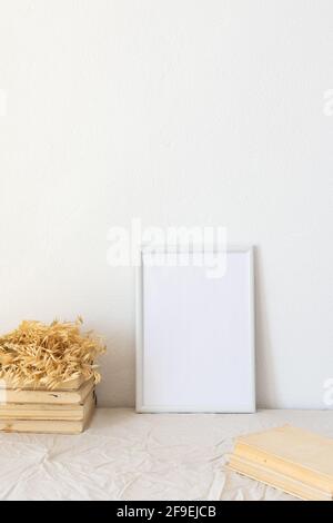 Wohnkultur-Modell, leerer Bilderrahmen in der Nähe von weiß lackierter Betonwand, getrockneten gelben Haferhalmen und alten Büchern Stockfoto