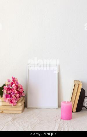 Heimdekor-Modell, leerer Bilderrahmen in der Nähe einer weiß gestrichenen Betonwand, ein Zweig rosa Hortensien auf alten Büchern und eine Kerze Stockfoto