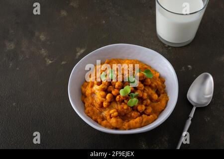 Dicke Kürbissuppe mit gebackenen Kichererbsen und Rettichsprossen auf einem Teller mit einem Glas Milch auf dem Tisch, leckeres gesundes Abendessen Stockfoto