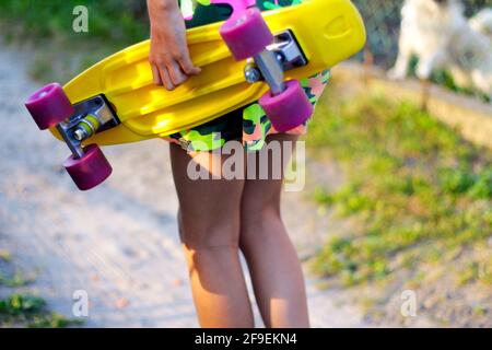 Defokussieren Mädchen mit gelben Penny Board. Blick von hinten. Sommer in leuchtenden Farben. Kinderhände halten tagsüber ein kurzes Cruiser-Pennyboard im Freien. Wellness Stockfoto