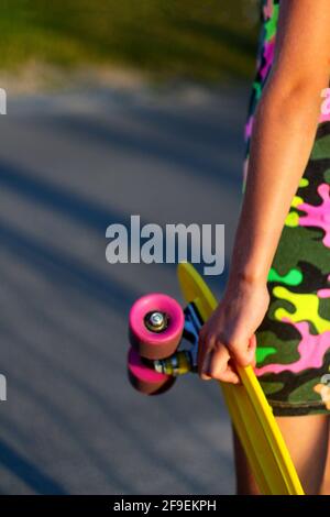 Defokussieren Mädchen mit gelben Penny Board. Rückansicht. Jugend-Hipster-Kultur. Nahaufnahmen von Kinderhänden, die tagsüber ein kurzes Cruiser-Pennyboard im Freien halten. Acti Stockfoto