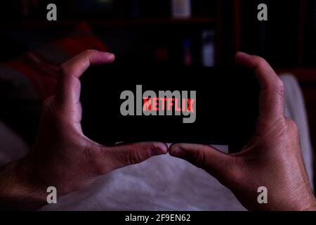 Spanien. 04, 18, 2021. Nahaufnahme eines Mannes, der nachts ein Smartphone mit dem Netflix-Startbildschirm auf seinem Sofa hält Stockfoto