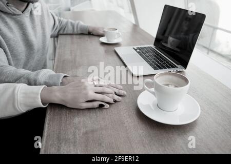 Frauenhand mit goldenem Ring bedeckt Männerhand. Romantisches Datumskonzept. Moderner Laptop und zwei leere Kaffeetassen auf Holztisch. Lässiger Style Stockfoto