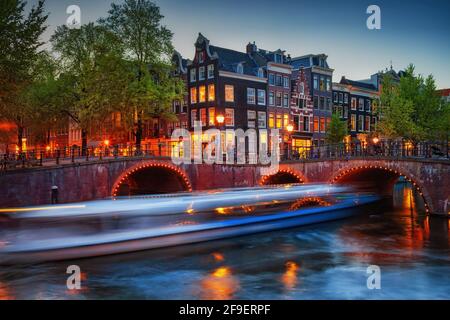 Abenddämmerung in der Stadt Amsterdam, Brücken auf Keizersgracht und Leliegracht Kanäle und verschwommene Form eines Kreuzfahrtschiffes, Nordholland, Niederlande. Stockfoto