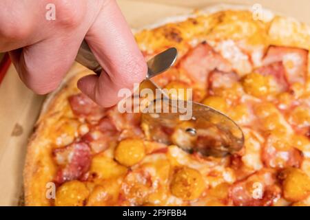 Nahaufnahme einer Hand mit einem Pizzaschneider Eine Pizza mit Speck und Huhn knallt auf eine Schachtel Stockfoto