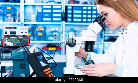 Eine junge Frau testet ein elektronisches Modul für die Produktion von Elektroautos. Qualitätskontrolllabor mit Mikroskop und Messgerät Stockfoto