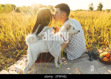 Stilvolles Paar küsst, entspannt in sonnigem Licht mit weißem Hund auf Decke zwischen Gras in Sommerwiese. Sommerurlaub und Picknick. Junge Familie enjo Stockfoto