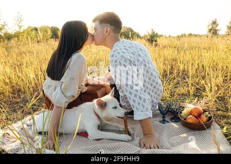 Stilvolles Paar küsst, entspannt in sonnigem Licht mit weißem Hund auf Decke zwischen Gras in Sommerwiese. Sommerurlaub und Picknick. Junge Familie enjo Stockfoto