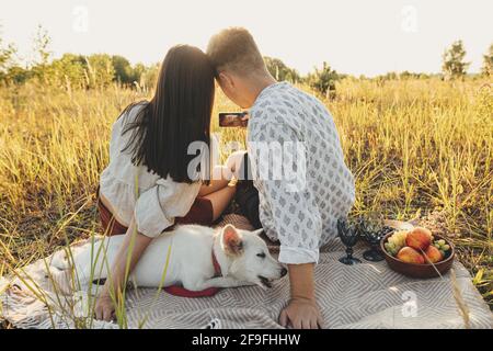 Stilvolles Paar, das Selfie-Fotos am Telefon fotografiert, während es sich mit einem weißen Hund auf der Sommerwiese unter Gras entspannt. Sommerurlaub und Picknick. Junge Familie genießen Stockfoto