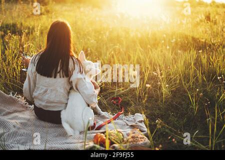 Stilvolle Frau genießen Sonnenuntergang mit weißem Hund auf Decke in warmen sonnigen Licht auf Sommerwiese. Urlaub und Picknick mit Haustier. Junge Boho weiblich entspannend Stockfoto