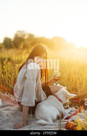 Stilvolle Frau, die sich mit ihrem weißen Hund auf der Decke im warmen, sonnigen Licht auf der Sommerwiese entspannt. Urlaub und Picknick mit Haustier. Junge Boho-Hündin genießt su Stockfoto