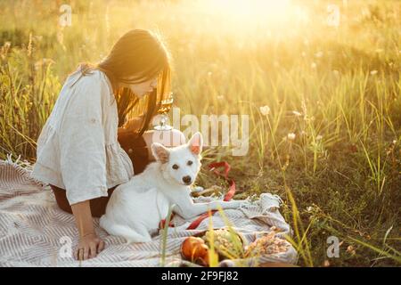 Stilvolle Frau, die sich mit ihrem weißen Hund auf der Decke im warmen, sonnigen Licht auf der Sommerwiese entspannt. Urlaub und Picknick mit Haustier. Junge Boho-Hündin genießt su Stockfoto