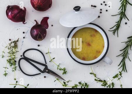 Draufsicht auf Kartoffelsuppe in weißer Schüssel mit Griff und Deckel auf weißem Hintergrund, verziert mit roten Zwiebeln, Vintage-Schere und Kräutern Stockfoto