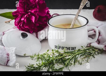 Kartoffelsuppe in weißer Schüssel mit Griff und Deckel auf weißem Tisch, verziert mit roten Zwiebeln, violetter Pfingstrose und Kräutern. Stockfoto