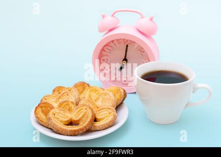 Weiße Tasse dampfenden heißen schwarzen Kaffee oder heiße Schokolade, frisch gebackene herzförmige Kekse und rosa Wecker auf hellblauem Hintergrund, Kopierraum. Stockfoto