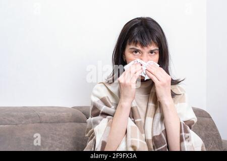 Krankheitstag zu Hause. Eine kranke Frau, die auf dem Sofa sitzt und ein Taschentuch hält, das ihre Nase bläst, fühlt sich ungesund an. Saisonale Krankheit, Allergie, Grippe-Konzept Stockfoto