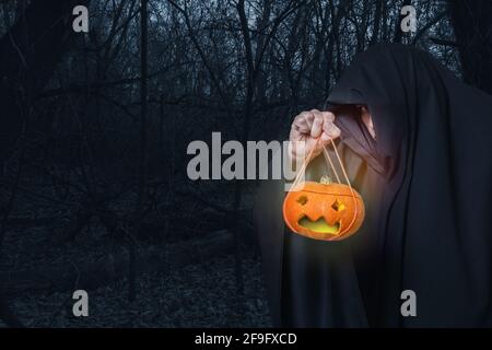 Ein Mann in schwarzer Kleidung hält eine Jack-o'-Laterne, die Laterne leuchtet mit gelbem Licht in einem gruseligen Nachtwald, Halloween-Hintergrund, Kopierraum Stockfoto