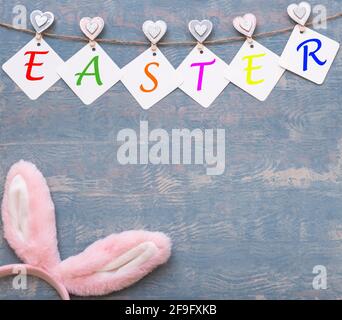 Ostern bunte Karte aus Girlande mit OSTERAUFSCHRIFT und Ostern rosa Hasenohren auf blauem Holzhintergrund, Kopierraum, flaches Lay, DIY. Frohes Ea Stockfoto