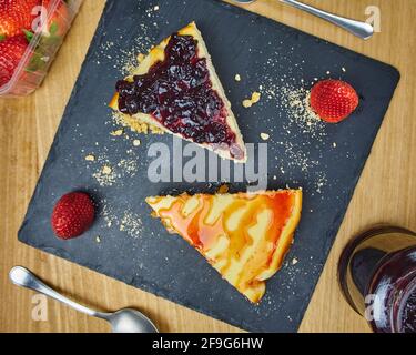 Set von Käsekuchen mit Erdbeer, Erdbeersoße, zerbröckeltem Gebäck und zwei Erdbeeren auf einem schwarzen Schieferbrett. Draufsicht Stockfoto