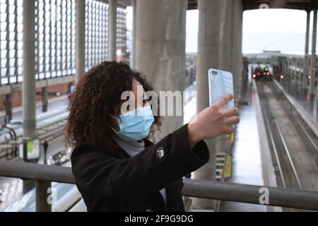 Porträt einer jungen Frau mit einer Maske, die im Hintergrund ein Selfie mit einem Smartphone an einem Bahnhof gemacht hat. Stockfoto