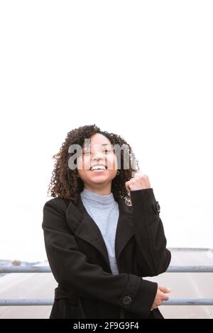 Lächelnde junge Frau mit lockigen Haaren, die direkt auf die Kamera schaut, mit weißem Hintergrund, in schwarzem Wollmantel gekleidet. Stockfoto